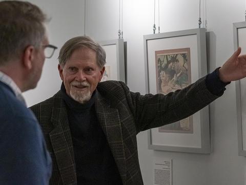 Arnold Satterwait, wearing a black turtleneck and dark jacket, gestured to a framed Japanese print in a gallery.