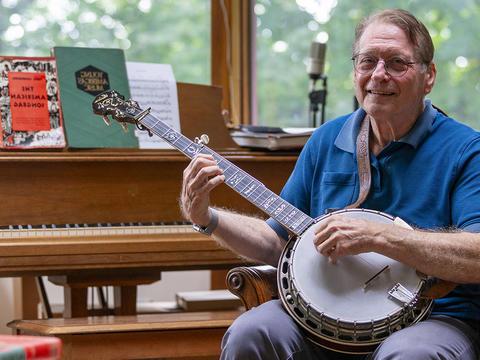 George Stavis holds a banjo and sits on a piano bench in front of a light brown piano.