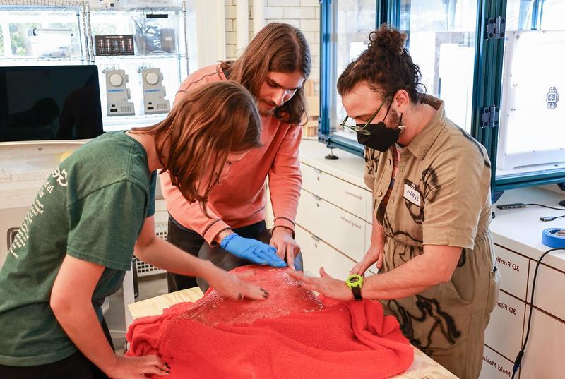Three people stand around a red piece of cloth and use their hands to shape a piece of alternative leather.