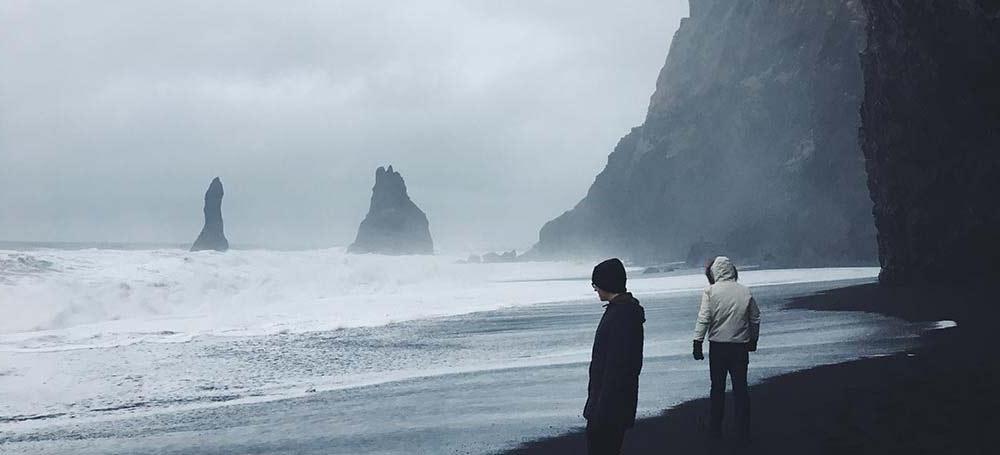 Two figures bundled in winter coats wander a black sand beach with the ocean roaring in the background